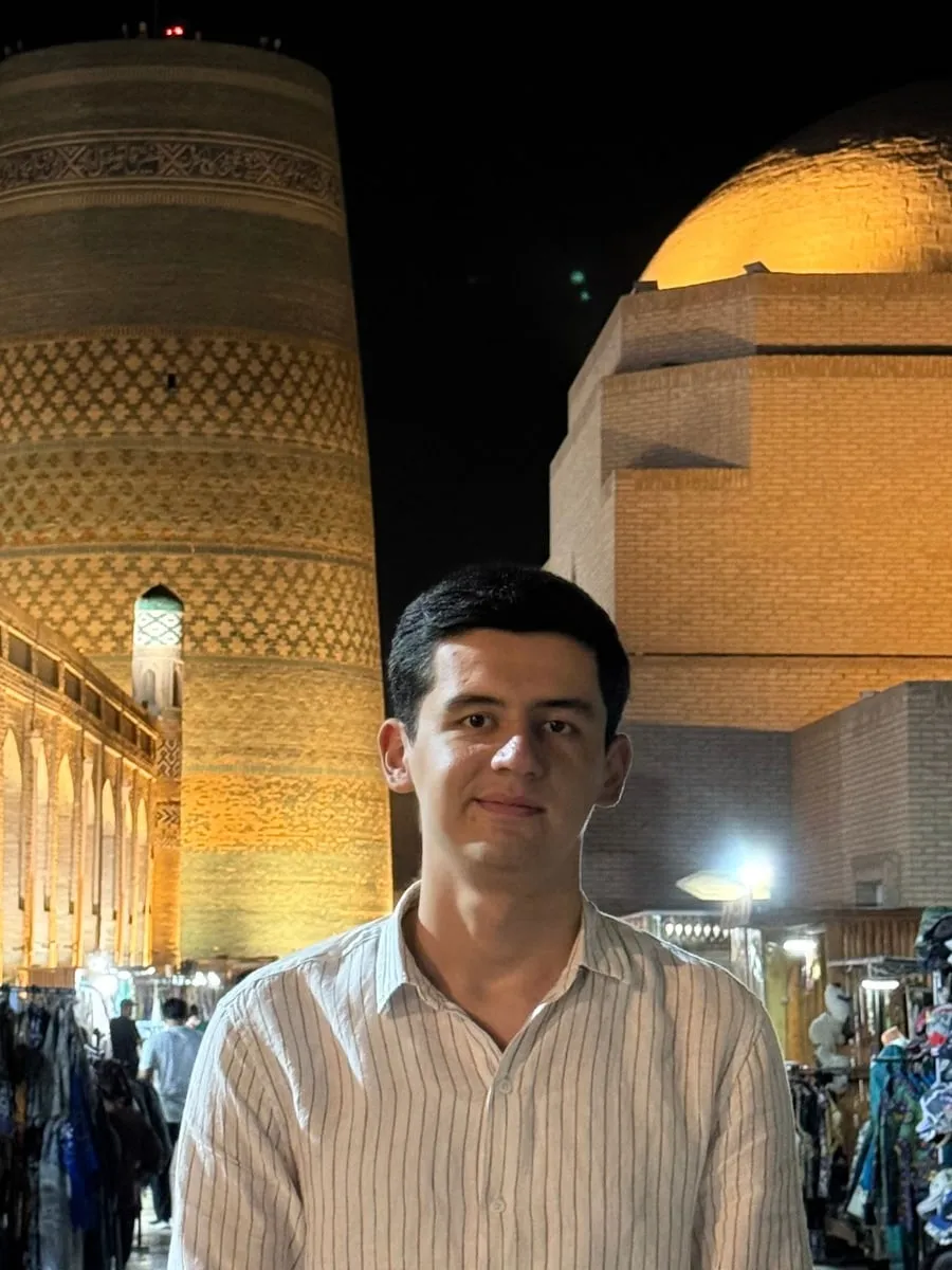 Asrorbek standing in Khiva's Itchan Kala at night, the Kalta Minor minaret lit up behind him